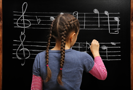Cute girl writing at the blackboard with musical notes, in the classroom