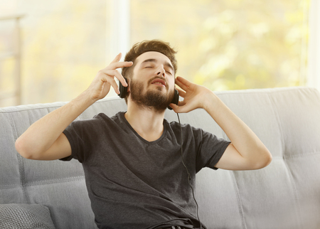 Handsome man emotionally listens music with headphones on grey sofa in the roomの写真素材