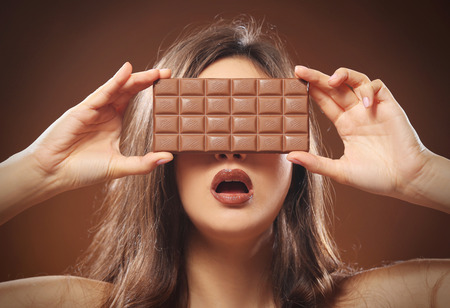 Portrait of young brunette closing her eyes with chocolate bar on brown background, close upの写真素材