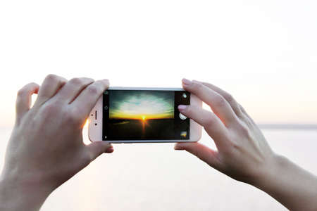 Female hands holding smart phone and taking photo of sunsetの写真素材
