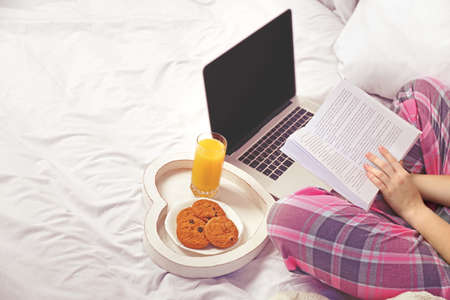 Woman in pajamas with laptop reading a book on her bedの写真素材