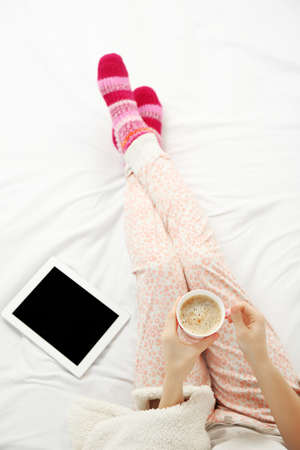 Woman in pajamas with tablet drinking coffee on her bed