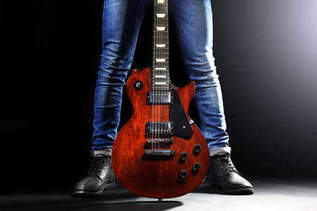 Young man in blue jeans standing with electric guitar on lighted dark background