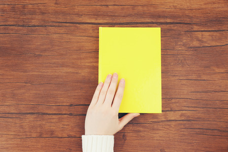 Female hand holding a yellow book cover on the wooden desk, top viewの写真素材
