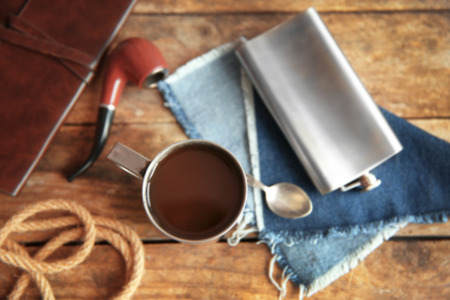 Metal mug of coffee with flask, spoon and tobacco pipe on wooden background