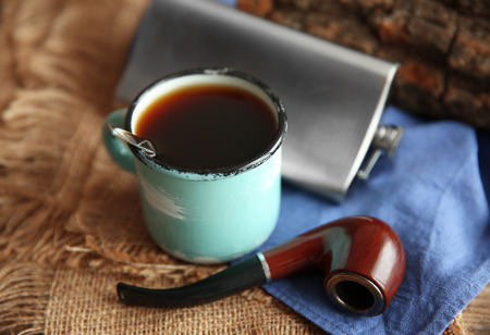Metal mug of coffee with flask and tobacco pipe on wooden background