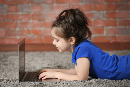 Little girl using laptop on fur carpet against brick wall background