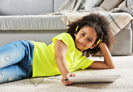 Afro-American little girl with headphones and tablet lying on floorの写真素材