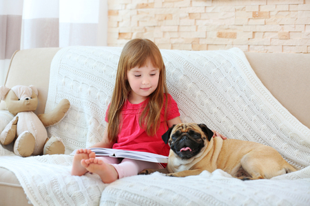 Adorable little girl reading book with cute pug on bedの写真素材