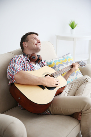 Young man with headphones playing guitar and sitting on a couch in light roomの写真素材