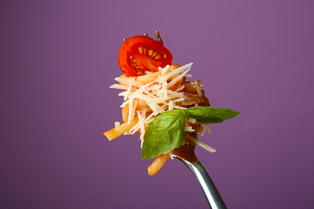 Fork with tasty pasta, cheese, tomato and basil on color background, close up viewの写真素材