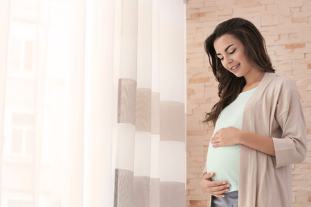 Beautiful pregnant woman standing near window at home