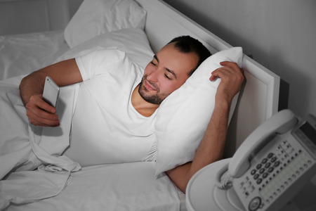 Young man with smart phone lying in bedの写真素材