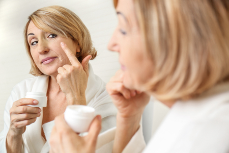 Senior woman applying cosmetic cream on face near mirrorの写真素材
