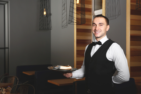 Young handsome waiter holding plate with delicious cake in a restaurantの写真素材