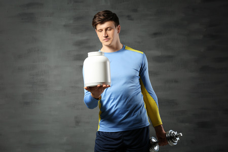 Young handsome sportsman holding bottle of protein and dumbbell on color backgroundの写真素材