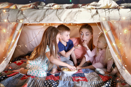 Young woman and cute children eating popcorn in hovel at homeの写真素材