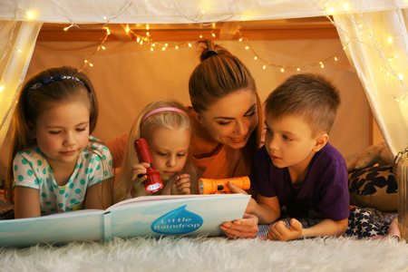 Young woman and cute children reading book in hovel at homeの写真素材