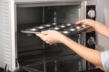 Young woman putting muffin tray into oven in kitchen