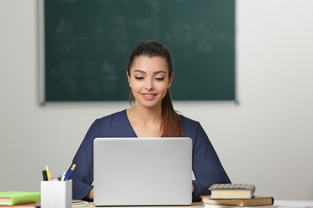 Beautiful young teacher working with laptop while sitting at table in classroomの写真素材