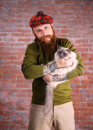 Young bearded man in plaid cap with fluffy cat on brick wall backgroundの写真素材