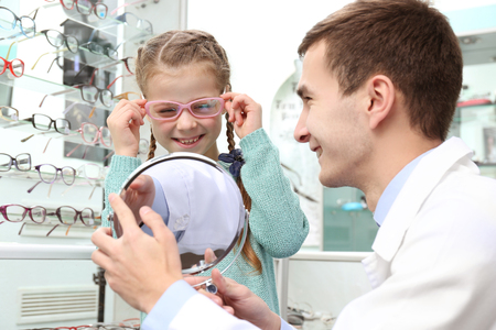 Young doctor helping little girl to choose new glasses in shopの写真素材