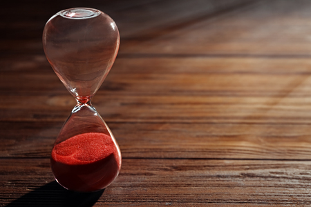 Time passing concept. Crystal hourglass with red sand on wooden background