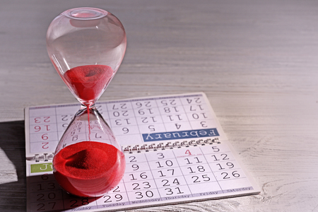 Time passing concept. Crystal hourglass with red sand and calendar on light wooden background
