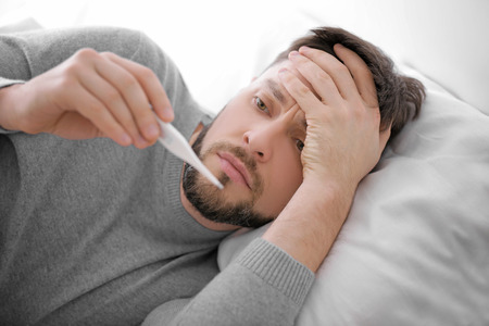 Young ill man measuring temperature while lying in bed at home