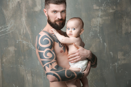 Handsome tattooed young man holding cute little baby on gray backgroundの写真素材