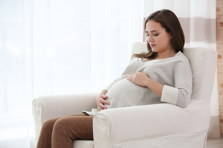 Beautiful pregnant woman sitting in arm-chair at homeの写真素材
