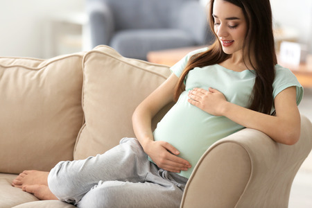 Beautiful pregnant woman sitting on sofa at homeの写真素材