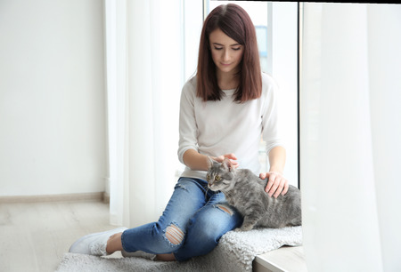 Beautiful young woman with cute cat sitting on window sill at homeの写真素材