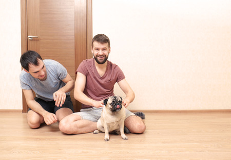 Happy gay couple sitting on floor and playing with pug dog at homeの写真素材