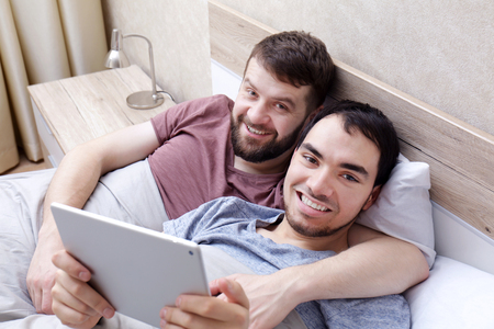 Gay couple lying on bed and using tablet at homeの写真素材