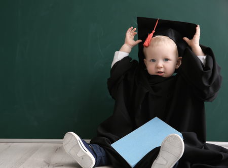 Cute little boy with book, magister hat and gown sitting on floor near blackboardの写真素材