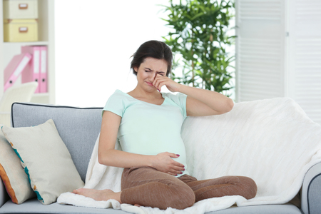 Young pregnant woman crying while sitting on sofa at homeの写真素材