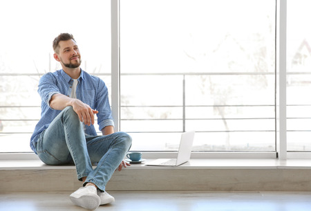 Happy young man sitting on windowsill with laptop and cup of coffeeの写真素材
