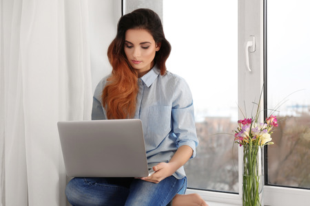 Beautiful young woman with laptop sitting on window sill at homeの写真素材