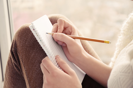 Beautiful young woman writing in notebook while sitting on window sill, closeupの写真素材