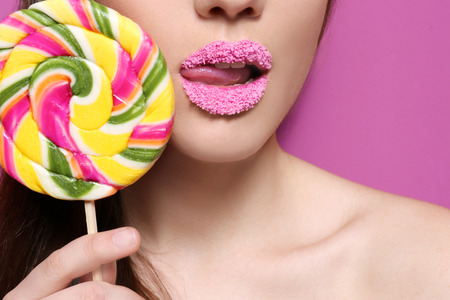 Beautiful young woman holding lollipop near lips covered with sugar on color background, closeup