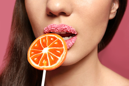 Beautiful young woman holding lollipop near lips covered with sugar on color background, closeup