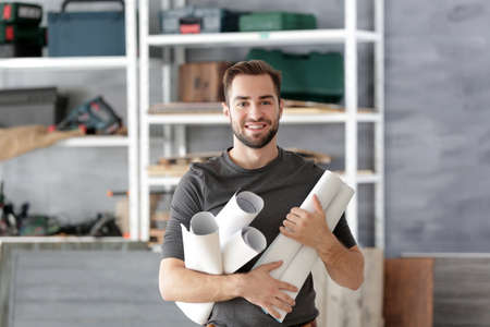 Young smiling carpenter holding paper rolls in light workshop