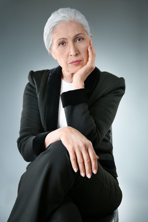 Beautiful elegant elderly woman sitting on chair against gray backgroundの写真素材