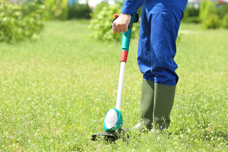 Young worker mowing lawn with grass trimmer outdoors on sunny day