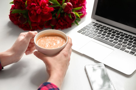 Female hands holding cup of coffee on table with computer, phone and peoniesの写真素材