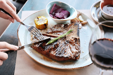 Woman's hands cutting tasty beefsteak on white plate with fork and knife on wooden tableの写真素材