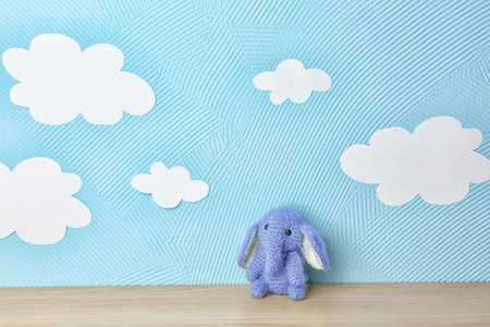Cute knitted toy elephant on wooden table near blue wall with paper cloudsの写真素材