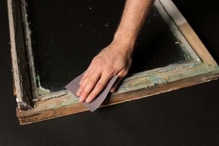 Hand of worker grinding surface of old window frame with abrasive paper, on dark background