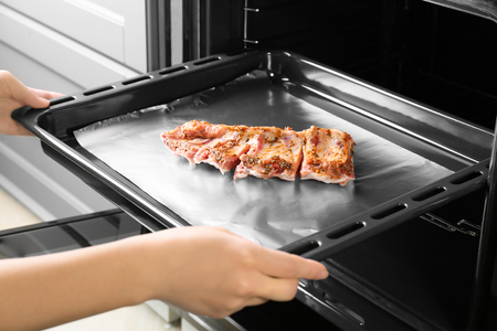 Young woman putting baking tray with pork ribs into ovenの写真素材